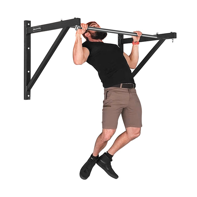 A person in a black t-shirt and brown shorts uses the Bells of Steel Adjustable Wall or Ceiling Mounted Pull Up Bar to perform a pull-up. They reach the pinnacle of their movement, with legs slightly bent and head tilted back.