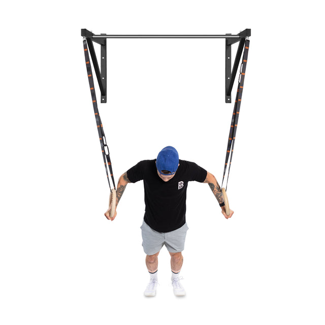 A person in a blue cap performs a chest exercise with gymnastic rings on the Bells of Steel Adjustable Wall or Ceiling Mounted Pull Up Bar. They wear a black t-shirt, gray shorts, and white shoes, maintaining a semi-upright position.