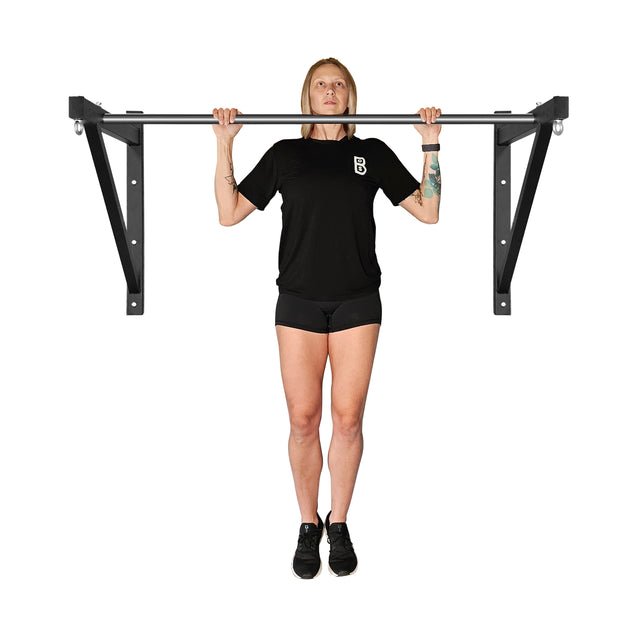 A person in a black t-shirt and shorts performs a pull-up using the Bells of Steel Adjustable Wall or Ceiling Mounted Pull Up Bar. Tattoos adorn both arms as they gaze straight ahead, framed against a plain white background.