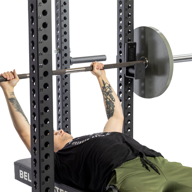 A person is performing a bench press on a weightlifting bench inside a power cage, equipped with Bells of Steel Roller J-Cups featuring durable nylon rollers. They hold a barbell, wearing a black T-shirt and green pants.