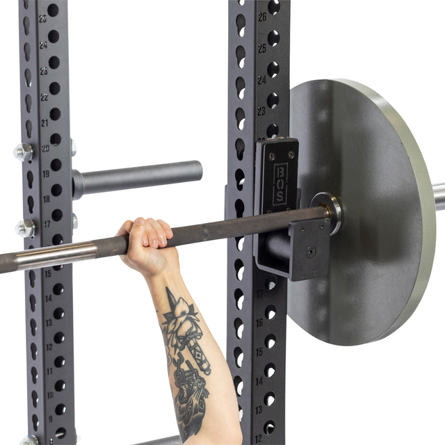 A person's tattooed arm, showcasing a striking animal design, grips a barbell with bench press accessories on a gym rack. Bells of Steel's Roller J-Cups cradle the weights securely, and the durable nylon rollers glide smoothly over the rugged industrial metal with numbered notches.