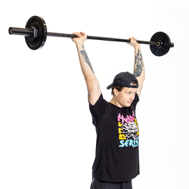 With tattooed arms, someone effortlessly lifts a Bells of Steel Short Rackable Barbell against a plain white background, showcasing their strength. The bar's durable black zinc coating complements their bold look, making it perfect for any compact home gym.