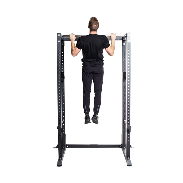 Dressed in black, a person effortlessly performs a pull-up on a bar attached to the Bells of Steel Residential Power Rack (2.3" x 2.3", ⅝" HOLES), effectively pulling their body upwards with their feet off the ground. The sturdy frame suggests an impressive home gym setup, ideal for reaching fitness goals.