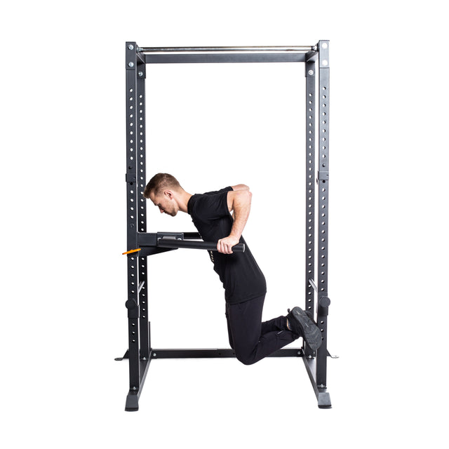 A man in black workout clothes does dips on parallel bars attached to a Bells of Steel Residential Power Rack (2.3" x 2.3", ⅝" holes) in his home gym, with his knees bent and feet off the ground.