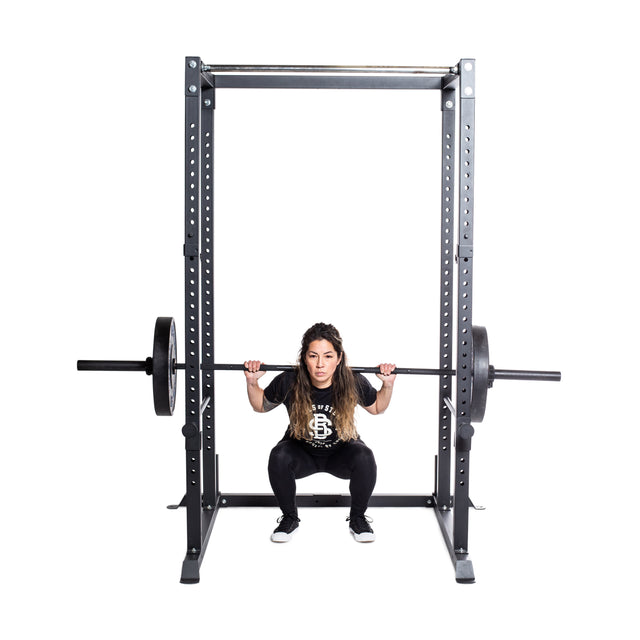 A person is executing a squat with a barbell on their shoulders, utilizing a Residential Power Rack (2.3" x 2.3", ⅝" HOLES) from Bells of Steel in their home gym. They maintain focus and proper squat form while wearing a black T-shirt and black shoes against a plain white background.