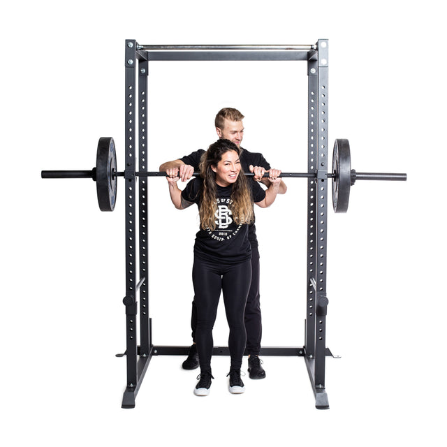 A woman is performing a squat using the Bells of Steel Residential Power Rack, with the barbell safely positioned on j-cups, while a man stands behind her to spot. Both are dressed in black athletic attire against a plain white backdrop, showcasing an ideal home gym environment.