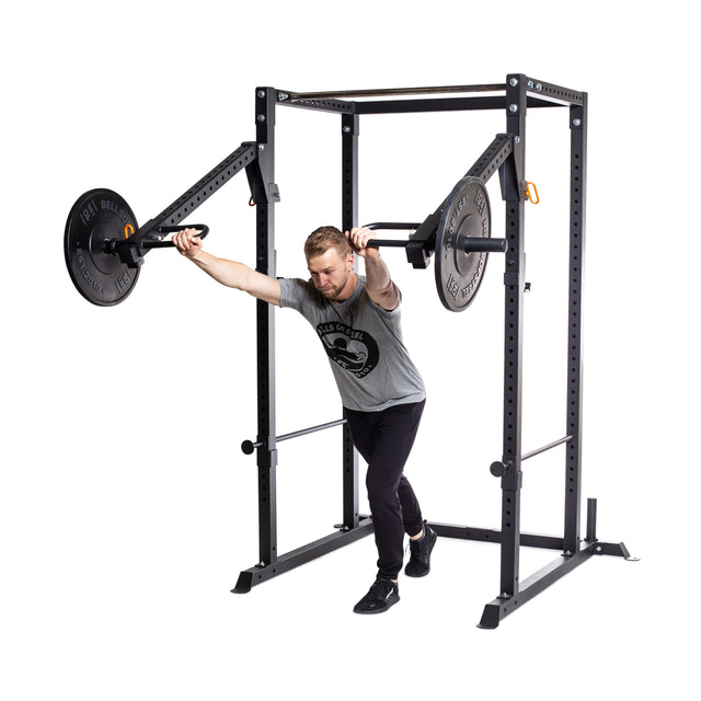 A man is performing a chest press exercise on the pivoting lever arm of the Bells of Steel Residential Power Rack in his home gym. He leans forward, gripping weighted bars while wearing a gray t-shirt and black pants. The elegant design of the Power Rack enhances the minimalist, white setting around him.