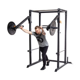 A man in athletic wear performs a chest press at home using the Bells of Steel Residential Power Rack (2.3" x 2.3", ⅝" holes) with j-cups and a barbell, leaning forward with arms extended inside the metal frame.