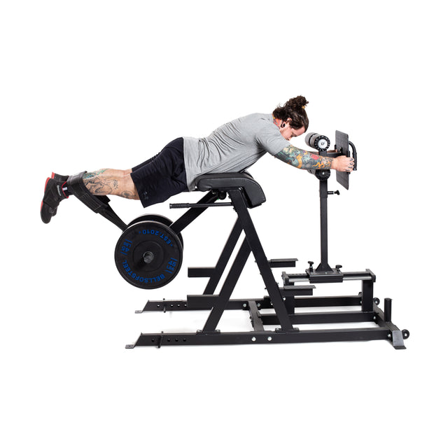 A person exercises on the Bells of Steel Reverse Hammer 2 In 1 GHD and Reverse Back Extension, focusing on the posterior chain. They are dressed in a gray shirt and black shorts, showcasing tattoos on their arms, while using the machine that has black padding and weights on its sides.