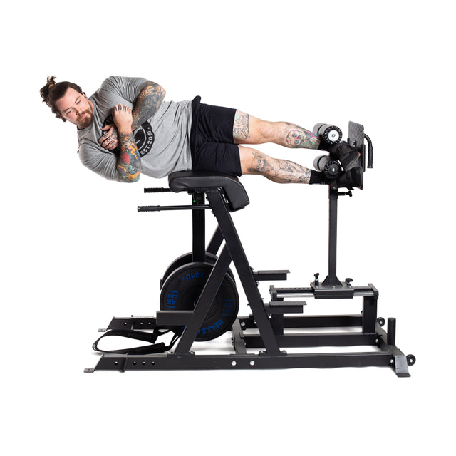 A tattooed individual is demonstrating a reverse back extension on the Reverse Hammer 2 In 1 GHD And Reverse Back Extension by Bells of Steel. Lying on their side, they are wearing a gray shirt and black shorts. This machine, featuring weights and cushioning, is designed to efficiently work the posterior chain.