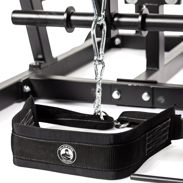 A close-up shows the Bells of Steel weightlifting belt with a chain attachment, placed on the Reverse Hammer 2 In 1 GHD And Reverse Back Extension equipment. This black belt, perfect for reverse back extensions, is highlighted by its white stitching and circular logo. Metal bars and bolts are integral to this fitness setup, engineered to enhance your posterior chain workout.