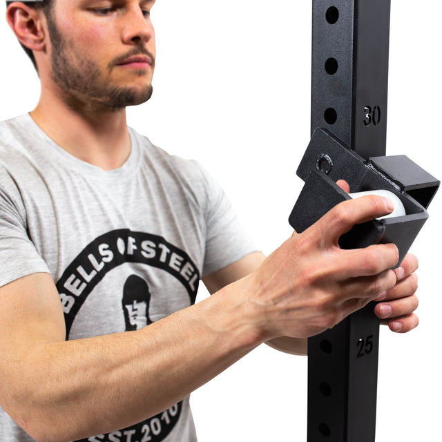 A person in a gray T-shirt featuring the "Bells of Steel" brand is adjusting a metal safety pin on a vertical gym rack equipped with Roller J-Cups. The rack, which includes height level markings, features durable nylon rollers ideal for bench press accessories.