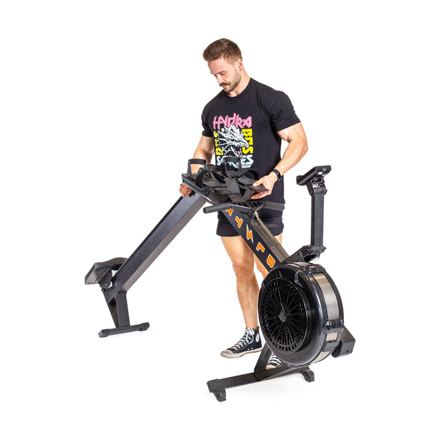 A person in a black T-shirt and shorts lifts the portable Blitz Air Rower by Bells of Steel. This foldable indoor rower, featuring an orange logo and large fan wheel, provides a low-impact cardio workout against a clear white background.