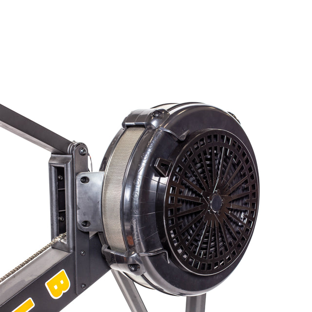 A detailed view of the fan mechanism on the Bells of Steel Blitz Air Rower, an indoor rowing machine. Enclosed in a black casing attached to the frame with visible chain, it's perfect for low-impact cardio, set against a plain white backdrop.