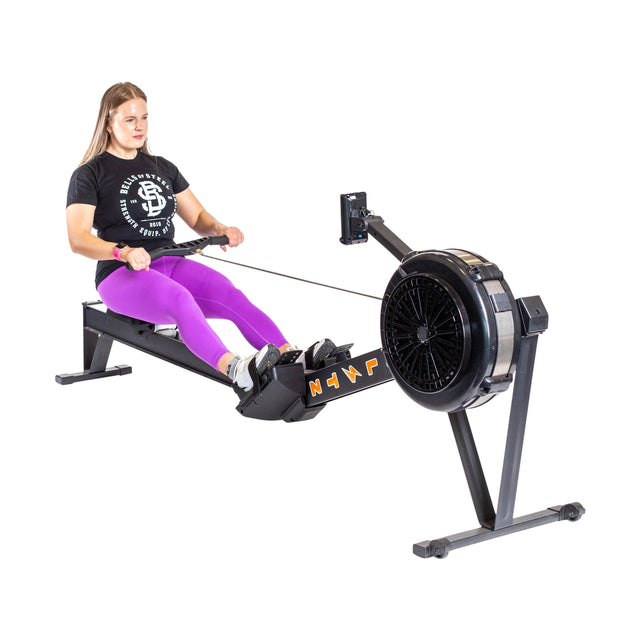 Wearing a black t-shirt and purple leggings, a person uses the Bells of Steel's Blitz Air Rower, an indoor low-impact cardio machine. They pull the handle with both hands while seated on the sliding seat, feet safely strapped in against a white background.