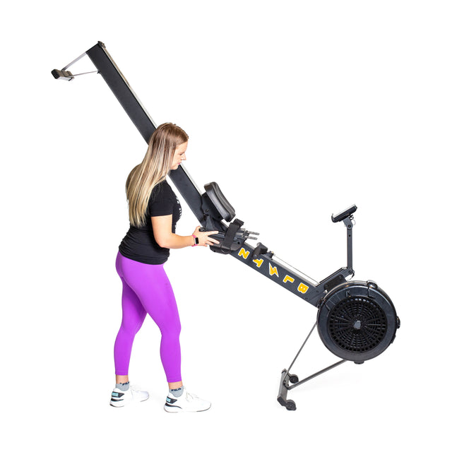 A woman in a black shirt and purple leggings tilts a Bells of Steel Blitz Air Rower onto its wheels. The sleek indoor rowing machine contrasts the plain white background, promising a low-impact cardio workout.