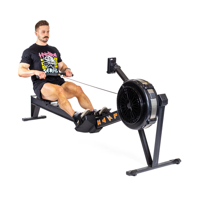 A man wearing a black t-shirt with colorful graphics and shorts is using the Bells of Steel Blitz Air Rower. He grips the handles with both hands, utilizing its fan wheel and digital display for a low-impact cardio workout indoors.