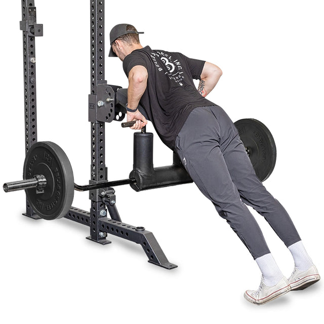 Wearing a black cap and shirt, a person focuses on their chest workout with a weight machine, leaning forward while gripping the handles. In gray pants, white socks, and sneakers, they exhibit solid form as if using Bells of Steel's Safety Squat Bar - The SS4 for versatile exercises.