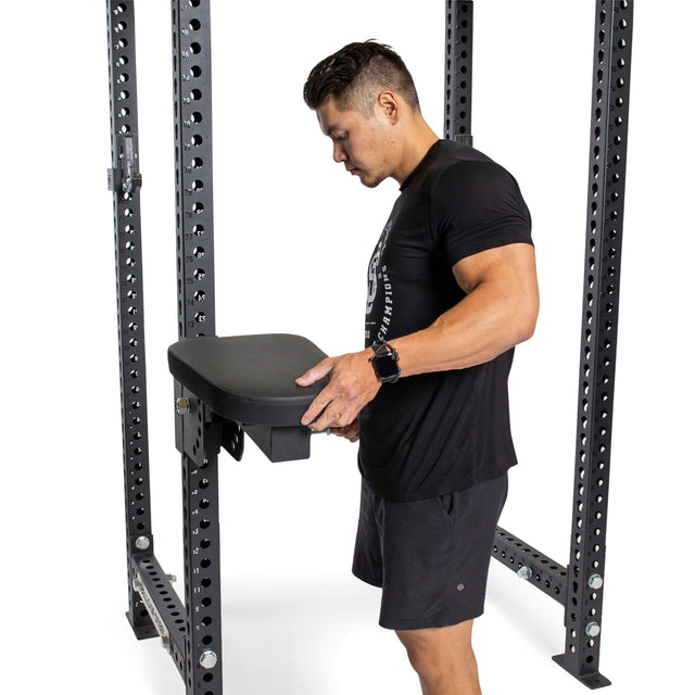 A man adjusts the Bells of Steel Seal Row Pad Rack Attachment. Dressed in a black T-shirt and shorts, he stands confidently within the metal frame. The plain white background highlights both the equipment and his focus on perfecting form.