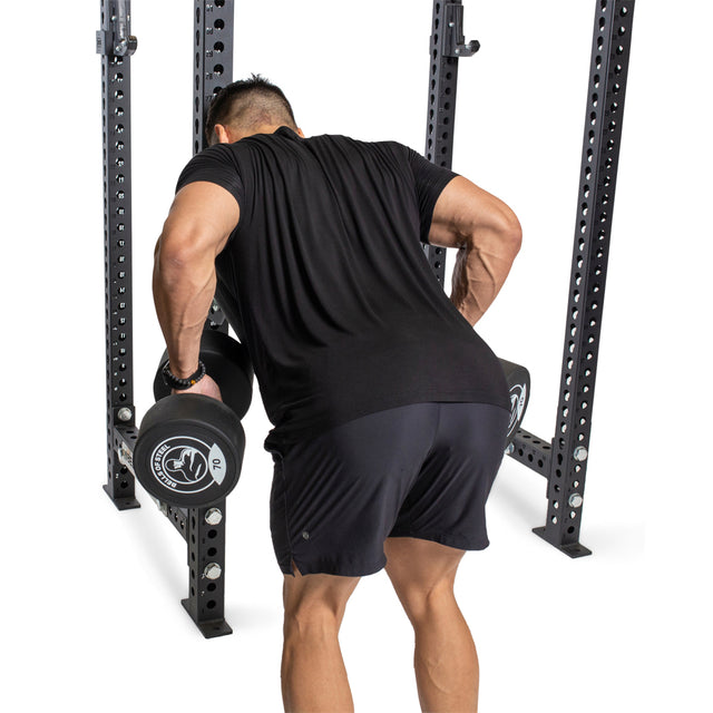 A man wearing a black t-shirt and shorts performs a bent-over row exercise with dumbbells in a gym setting, using the Bells of Steel Seal Row Pad Rack Attachment for added stability while standing in front of a power rack.