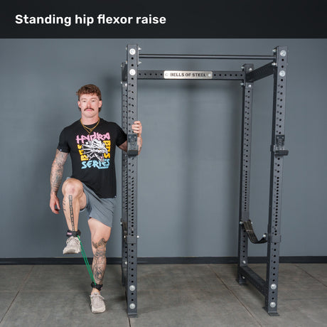 A man performs a standing hip flexor raise using the Bells of Steel Human Trainer - Leg Cords for ankle resistance, holding onto a squat rack for support as he lifts one knee while keeping his other foot on the ground.