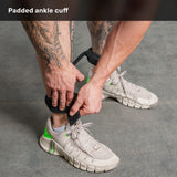 A person with tattoos puts on a padded ankle cuff over their sock while wearing light-colored sneakers with green accents, standing on a gray floor. They are preparing to use the Bells of Steel Human Trainer - Leg Cords for their workout, ensuring secure and comfortable support during leg exercises.