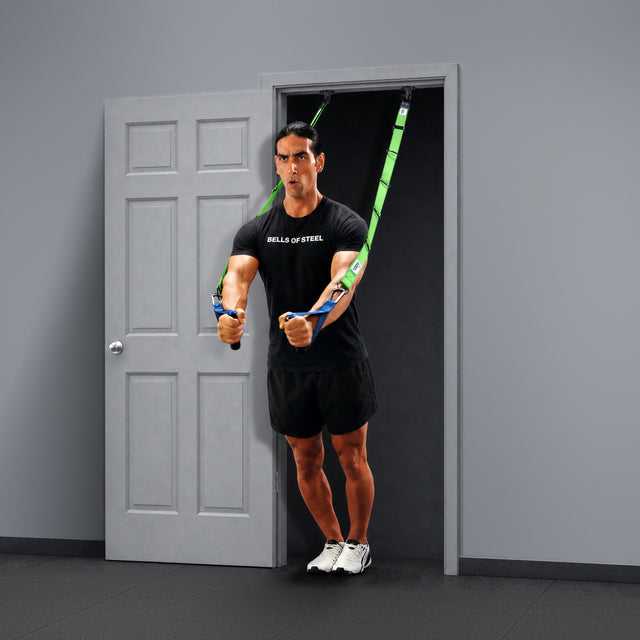 A person uses Bells of Steel Human Trainer - Ceiling Mounts attached to an anchor point for a workout, standing with arms extended forward and feet together, wearing a black “BELLS OF STEEL” shirt and shorts.