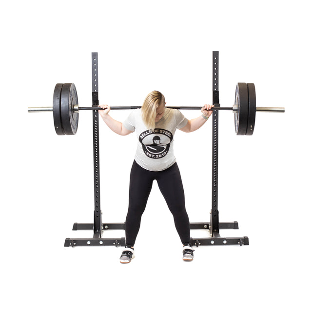 A person in a white t-shirt and black leggings performs a squat with a barbell on their shoulders, utilizing the Bells of Steel Squat Stands (2.3" x 2.3", ⅝" HOLES). The plain white background highlights the focus on weightlifting.