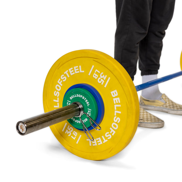 A person in black pants and checkered shoes stands next to a Bells of Steel Olympic barbell with yellow and green plates, secured by spring collars.