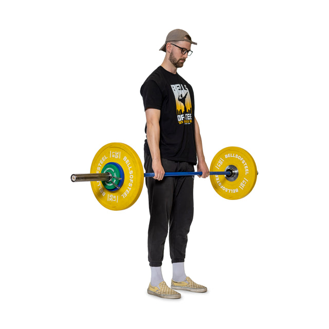 A man confidently holds an Olympic barbell with yellow plates, wearing a black shirt, dark pants, checkered shoes, a backward cap, and glasses. The vibrant Bells of Steel Spring Collars shine against the plain white background.