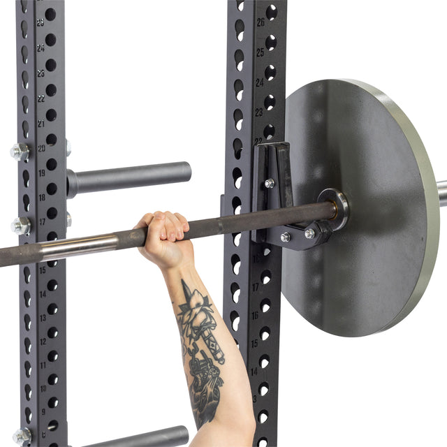 An arm with a tattoo grips a barbell loaded with weight plates on a squat rack, secured by Bells of Steel Sandwich J-Cups.