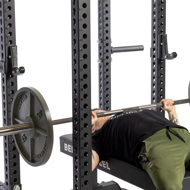 A person wearing a black shirt and green pants performs a bench press with heavy weights on a barbell, supported by the Bells of Steel Sandwich J-Cups on the power rack. The setup ensures stability, while the focus remains on the intense lifting activity.