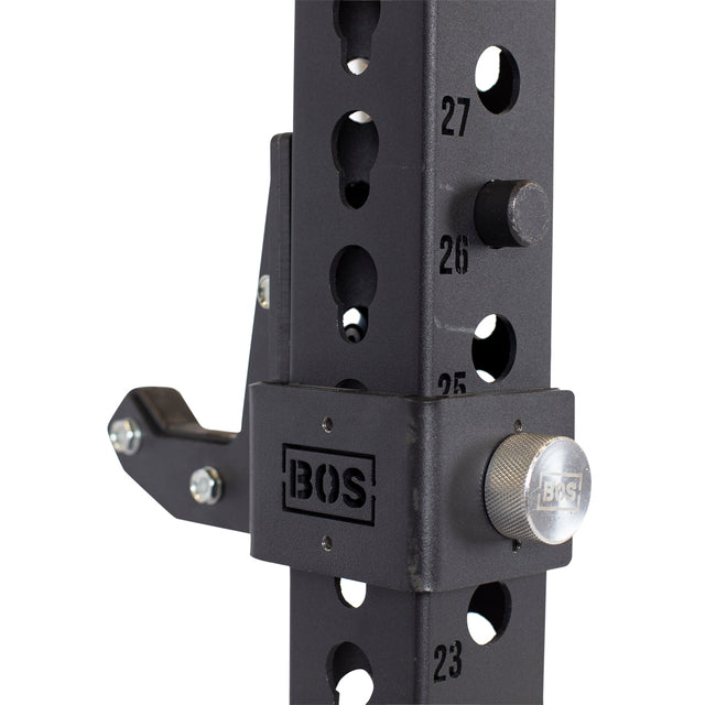 A close-up of a black metal gym rack shows numbered holes and adjustable hooks, highlighting the Sandwich J-Cups. The rack features a "Bells of Steel" branded plate and includes silver knobs and bolts for securing attachments. It also has HDPE padding to ensure durability and protection during workouts.