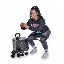 A woman in athletic wear uses the Bells of Steel Strength Circuit Starter Kit, squatting with a kettlebell near a box as dumbbells rest nearby—the perfect home workout setup against a white background.