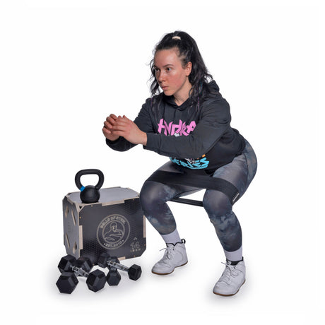 A woman in athletic wear uses the Bells of Steel Strength Circuit Starter Kit, squatting with a kettlebell near a box as dumbbells rest nearby—the perfect home workout setup against a white background.
