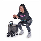 A woman with dark hair in a ponytail squats in front of a box of Bells of Steel Strength Circuit Starter Kits, surrounded by a kettlebell and dumbbells, ready for an energizing home workout on a white background.