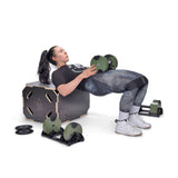 A woman in athletic wear does a barbell hip thrust with her upper back on a plyometric box. Bells of Steel Strength Circuit Starter Kits dumbbells and weight plates are nearby, ideal for an effective home workout.