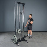A man stands by a tall, black cable machine in a gym with gray walls and a concrete floor, using Bells of Steel Fat Bar - Single D Handle attachments to enhance grip strength.