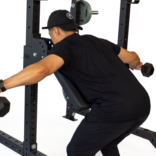 Dressed in a black outfit and cap, an individual is performing a bent-over row with dumbbells using the Seal Row Pad Rack Attachment by Bells of Steel for improved stability. Positioned within a squat rack, they display proper form and technique amidst various weightlifting equipment in the gym environment.