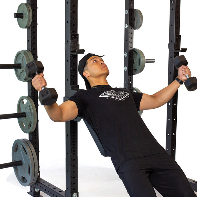 A person in a black T-shirt and cap demonstrates an incline dumbbell fly on a bench equipped with the Bells of Steel Seal Row Pad Rack Attachment for enhanced stability. In the background, weight racks and plates are visible, emphasizing the well-equipped gym atmosphere.