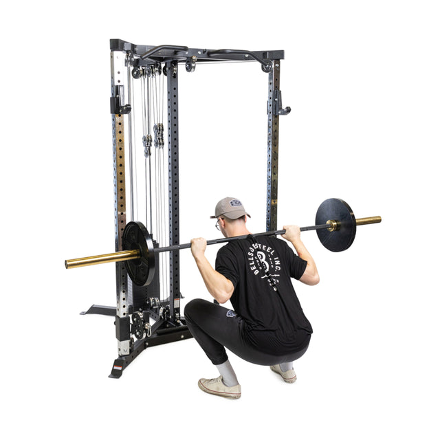 Dressed in black, someone performs a squat using the Bells of Steel All-in-One Trainer, featuring a barbell on a cable machine. The functional trainer system is set in a well-equipped gym with visible pulleys and cables.