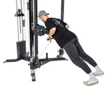 A person in a black shirt, cap, and glasses intently uses the Bells of Steel All-in-One Trainer in the gym, pushing against the bar of this versatile machine with adjustable weights and pulleys, ideal for a full-body workout.