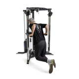 Wearing a black shirt, cap, and gray shoes, a person performs a tricep dip on the Bells of Steel All-in-One Trainer. Mid-rep with their back to the camera, they utilize the adjustable pulleys of the metallic gym equipment for an optimal workout.