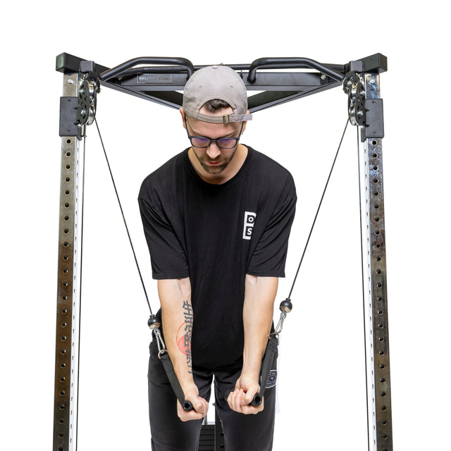 A person wearing a black shirt and gray cap utilizes the Bells of Steel All-in-One Trainer, grasping the handles with focus. The pulleys ensure a smooth workout experience on this functional trainer.