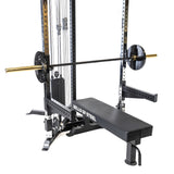 A Bells of Steel All-in-One Trainer on white flooring includes a black weight bench with an adjustable barbell rack, serving as a versatile squat stand, plus a barbell loaded with weights and a cable pulley system.