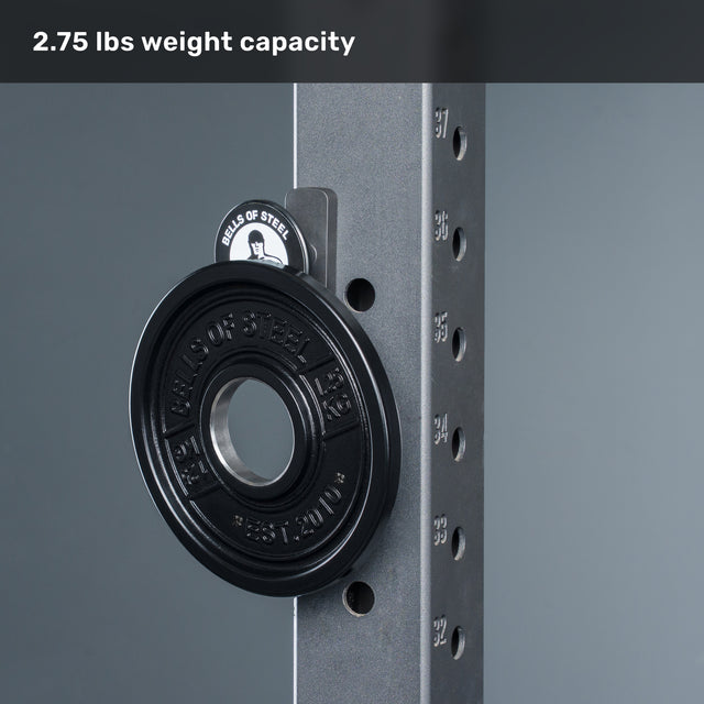 A black weight plate labeled “2.75 LBS” is mounted on a Bells of Steel metal rack with multiple holes, next to the Selfie Sentinel - Magnetic Rack Phone Holder. Text at the top reads “2.75 lbs weight capacity.” The background is plain gray.