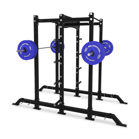 A black Hydra 2-Sided Rack from Bells of Steel, set up for squats with a barbell loaded with blue weight plates labeled "BELLOFSTEEL." This open ecosystem rack includes safety bars and hooks, providing modular components for versatile weightlifting exercises.