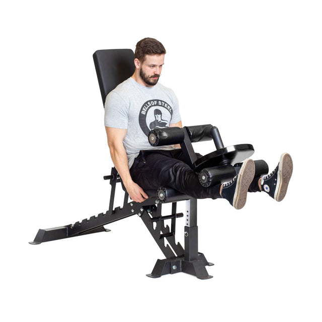 A man in a light gray t-shirt and black pants exercises by extending his legs forward while gripping the handles of the machine, seated on a Bells of Steel Buzz-Saw Heavy-Duty Adjustable Bench.