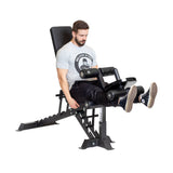 A man is seated on a Bells of Steel bench fitted with Bench Attachments, performing leg exercises. He wears a gray t-shirt and black pants, intently focusing on his workout using the adjustable bench's leg extension feature.