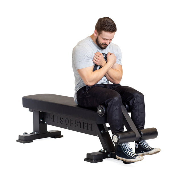 A man with a beard is seated on a Bells of Steel Bench Attachment, arms folded across his chest. He's dressed in a gray shirt, dark pants, and black sneakers. The bench proudly features adjustable components, with the brand name "Bells of Steel" prominently displayed on its side.